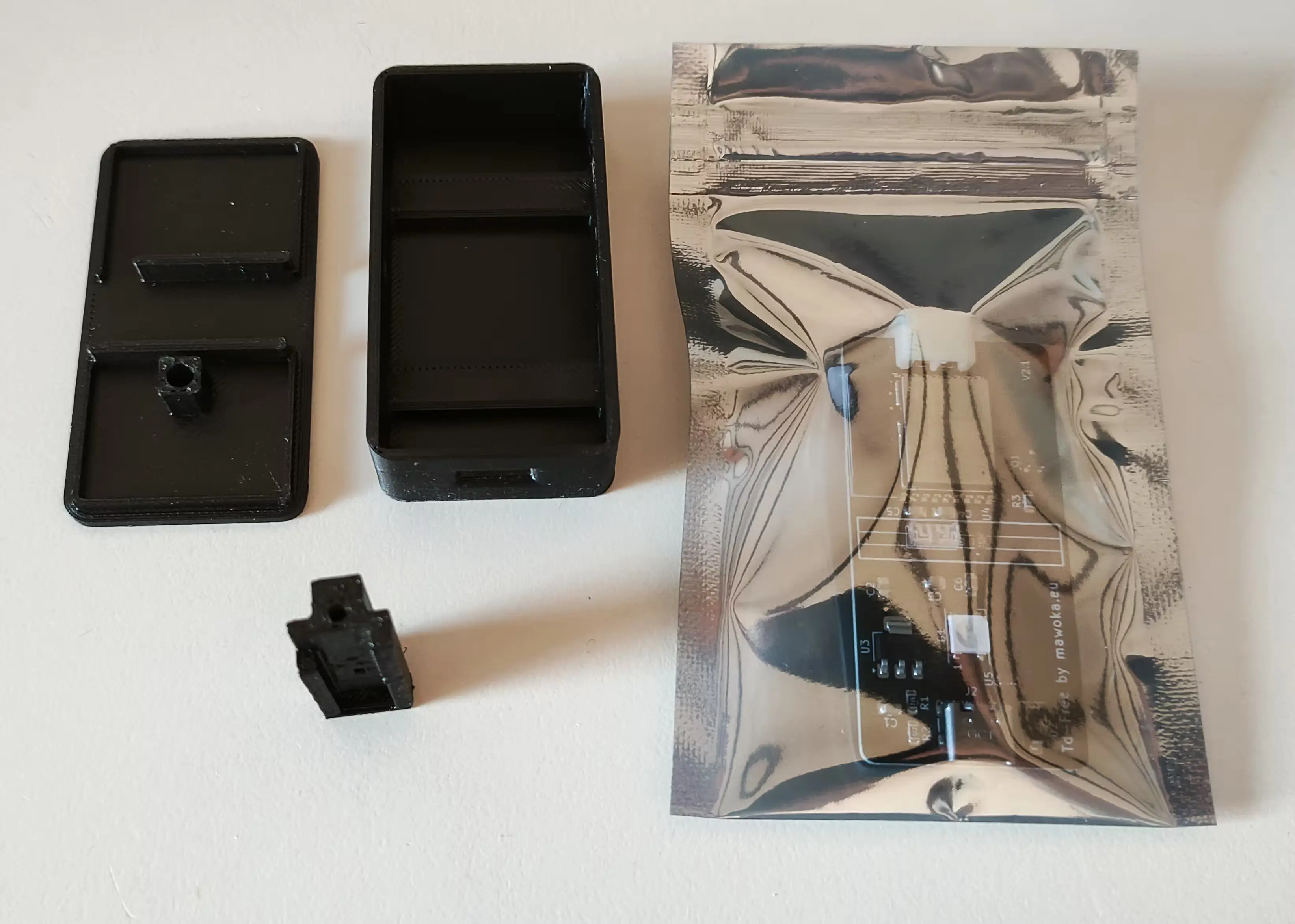 A black plastic electronics enclosure in three parts (lid, base, and a small internal component) alongside an anti-static bag containing a visible printed circuit board.