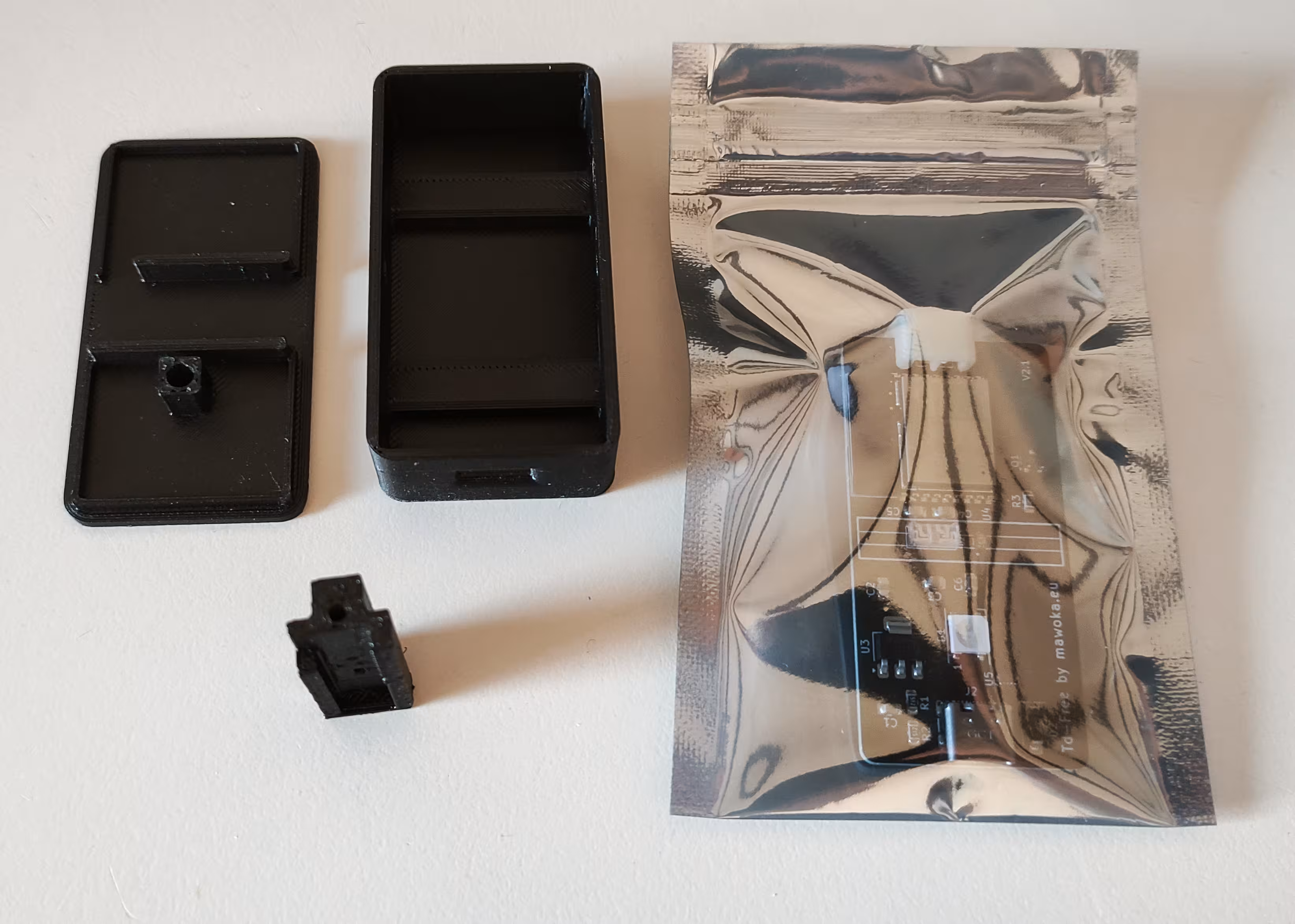 A black plastic electronics enclosure in three parts (lid, base, and a small internal component) alongside an anti-static bag containing a visible printed circuit board.